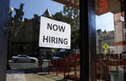 "Now hiring" sign on a storefront reflecting outdoor seating 