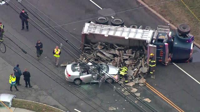 A dump truck lays on its side on a road next to a silver sedan with its roof cut off. Debris from the truck spills out onto the vehicle. 