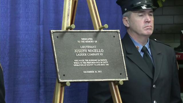 A member of the FDNY stands next to a plaque reading, "Dedicated to the memory of Lieutenant Joseph Maiello Ladder Company 83 who made the supreme sacrifice while in the performance of duty operating at Staten Island Box 1391. December 26, 2021." 