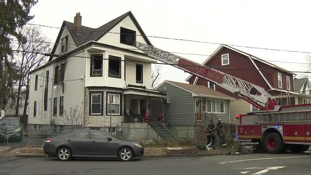 A ladder is extended from a fire truck to a window in the attic of a home with broken windows and visible smoke damage. 