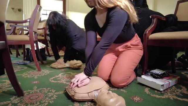 Two individuals kneel on the floor and practice CPR on training dummies. 