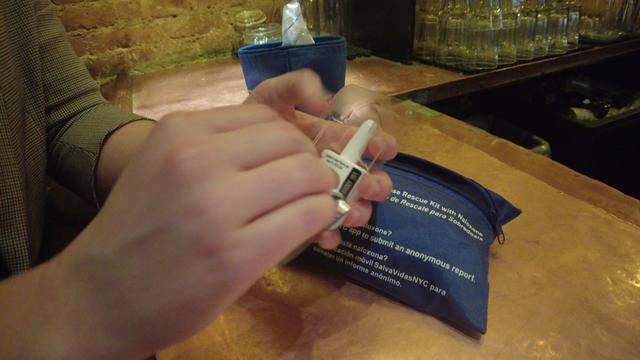 An individual sits at a bar holding a package of Narcan. 
