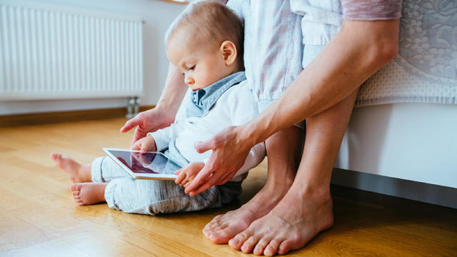 Cute infant baby boy barefoot using a tablet sitting on the flloor, his unrecognizable mother helping him to hold gadget. Real people life concept. 