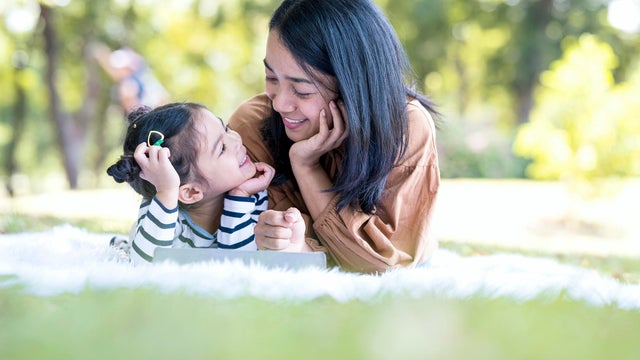 Asian Mother and cute daughter are looking each other while using digital tablet in the park on beautiful summer day. They are teaching and learning technology together. 