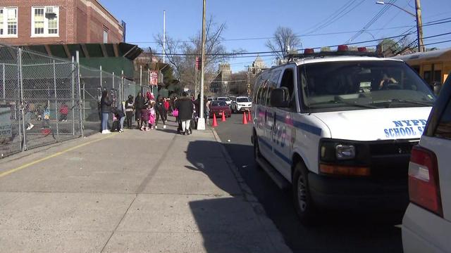 An NYPD school safety van sits parked outside an elementary school down the sidewalk from children and parents. 