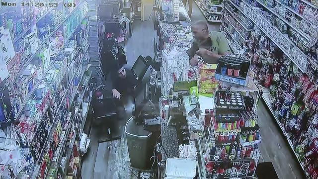 A man leans over the counter of a convenience store and points a rifle at an employee crouching on the floor. 