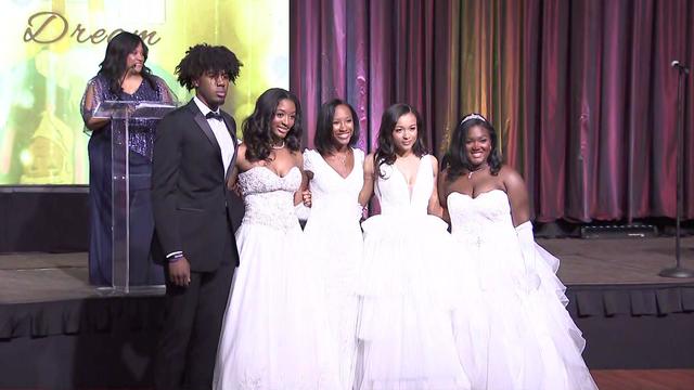 CBS2's Elise Finch stands on stage as teenagers in formal wear pose in front of her. 