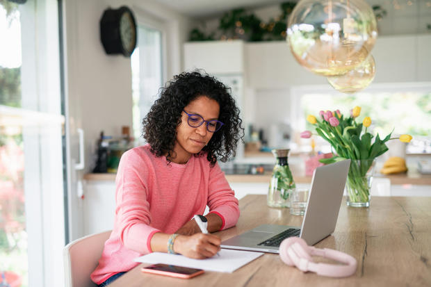 Multiracial woman in home office in kitchen, writing notes. 