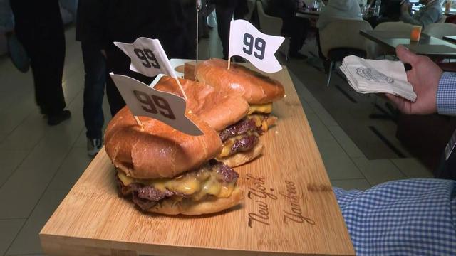 Three burgers with toothpicks that have white flags with the number 99 on them sit on a wooden cutting board that says "New York Yankees" on it. 