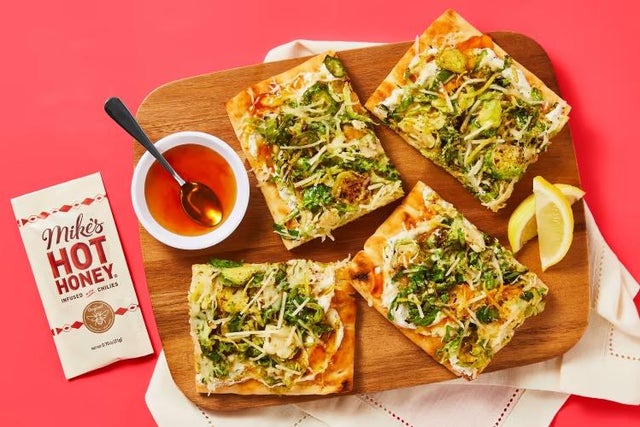 Hot honey brussels and ricotta flatbreads with parmesan and italian cheeses served on cutting board next to lemon wedges and a small bowl of hot honey. 