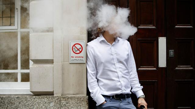 White smoke surrounds the head of a man leaning against a wall with "no smoking" sign 