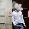 White smoke surrounds the head of a man leaning against a wall with "no smoking" sign 