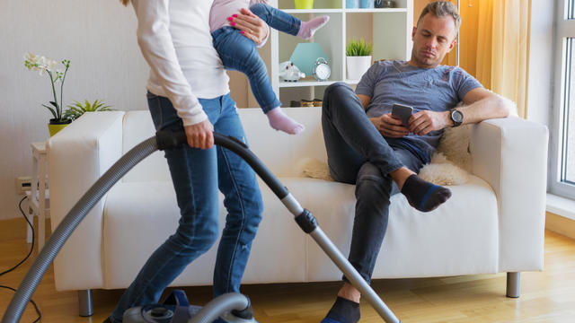 Woman with small child doing housekeeping while man sitting in couch 