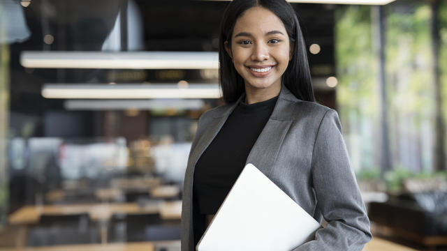 Smart People with Technologies to Boost Business Success. Portrait of Confident Young Female Entrepreneur standing while holding a laptop in a modern business office. 