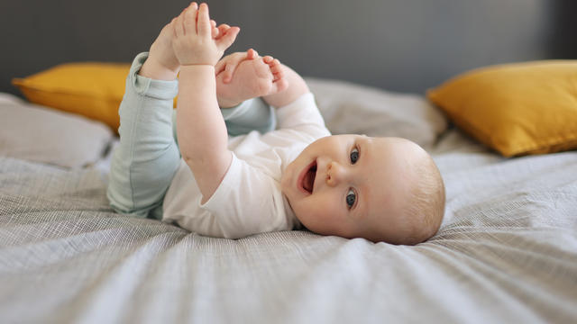 A 6 month old baby boy smiling, laying on a bed 