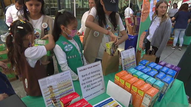 Girl Scouts stand behind a table full of Girl Scout cookies. 