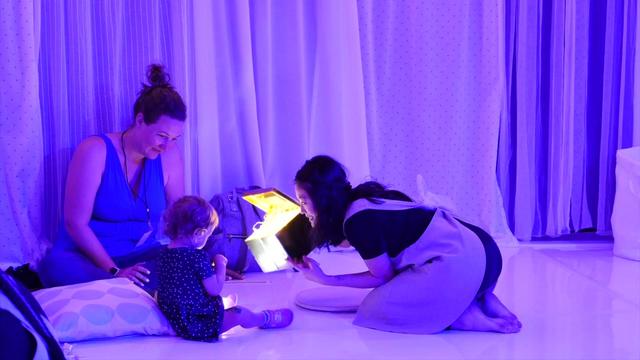 In a purple-lit room, a performer kneels on the floor and shows a young child a box with a light inside while a woman watches. 