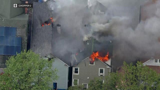 An aerial view of flames shooting from windows of the top floor of a home and breaking through the roof of a neighboring home. 