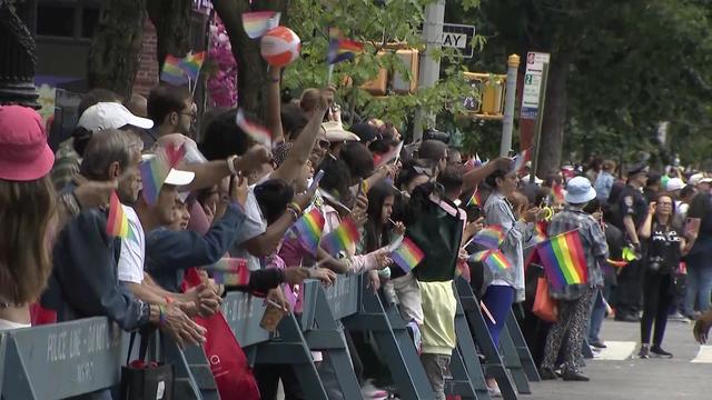 Spectators, many with rainbow flags, stand behind barricades along a Queens street. 