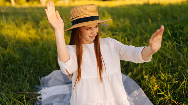 Portrait of dissatisfied redhead pretty woman in straw hat and white dress chasing insects away from herself sitting on field with green grass. 