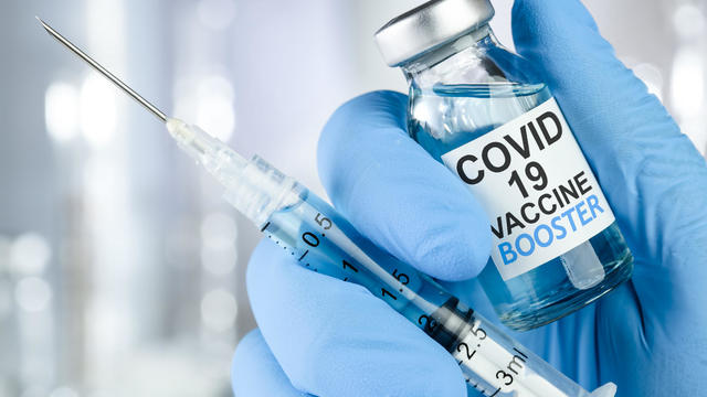 Hand in blue medical gloves holding a syringe and vaccine vial with Covid 19 Vaccine Booster text, for Coronavirus booster shot. 