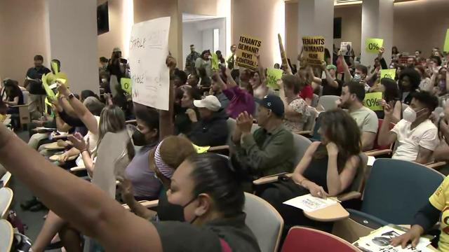 Tenants sit in the audience at a meeting, many holding signs reading "Tenants demand rent control!" and "0%." 