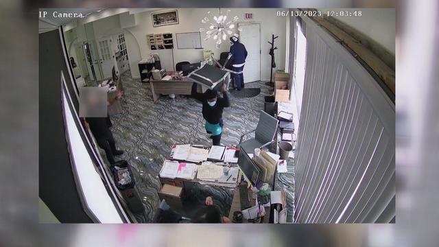 Surveillance video from inside an office shows a man holding an office chair above his head as a woman cowers behind a desk. Another man stands off to the side, and another suspect stands by a door in the back of the room. 