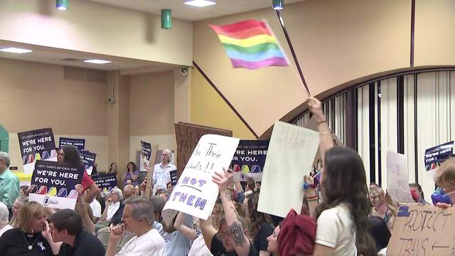 A crowd sits at a school board meeting, many holding signs reading "We're here for you." One individual waves a gay pride flag. 