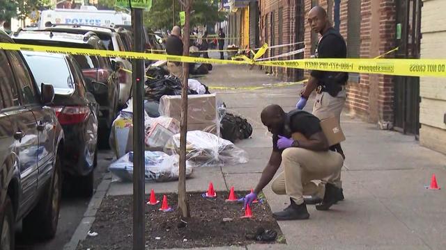 NYPD officers examine multiple small orange cones placed on a sidewalk behind crime scene tape. 