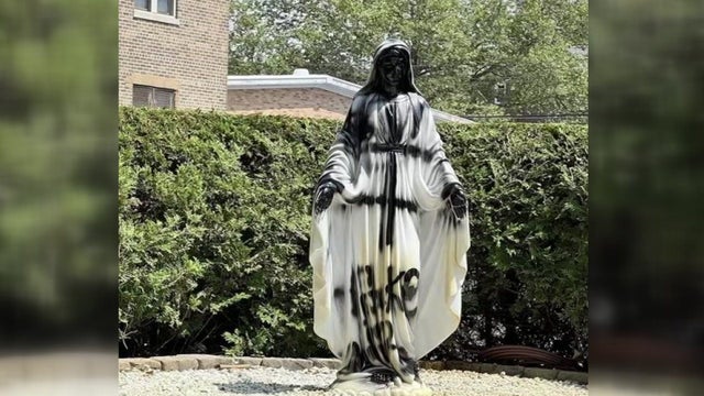 A white statue of the Virgin Mary that has black spray paint covering the fact, hands and feet and what appears to be the word "fake" on it. 