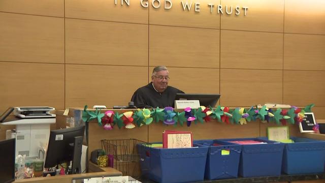 A judge sits in a courtroom that is decorated with a floral summer garland and colorful bins. 