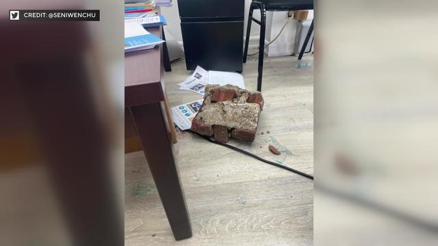 A photo shows three bricks cemented together laying on the floor of an office amid broken glass and papers. 