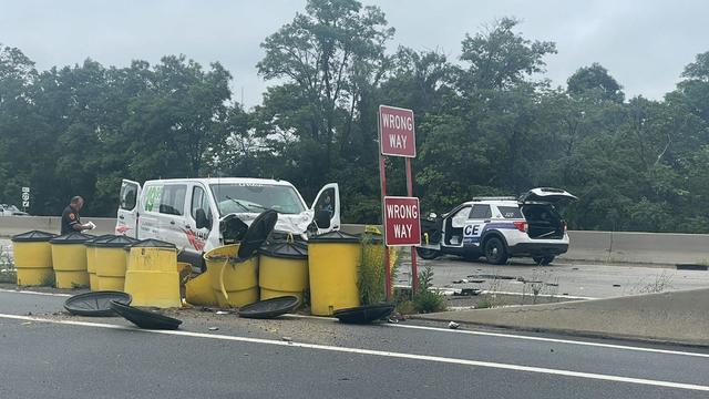 A U-Haul van is crashed into yellow barrels on a highway. Nearby a Suffolk County Police cruiser sits with its trunk and driver's side door open. 