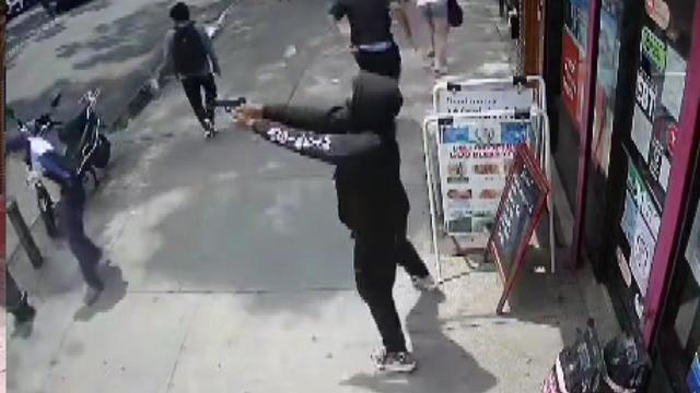 An individual standing on a sidewalk outside a store points a gun across the street. 