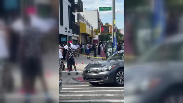 Crime scene tape blocks off a section of a sidewalk in Brooklyn while dozens of people stand nearby. 