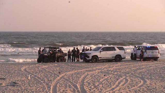 Three Long Beach police vehicles parked on a beach near the edge of the water as numerous police officers stand around. 