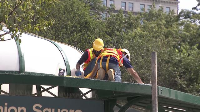Two individuals in hard hats and orange vests kneel on the roof of the Union Square subway station entrance as they clean it. 
