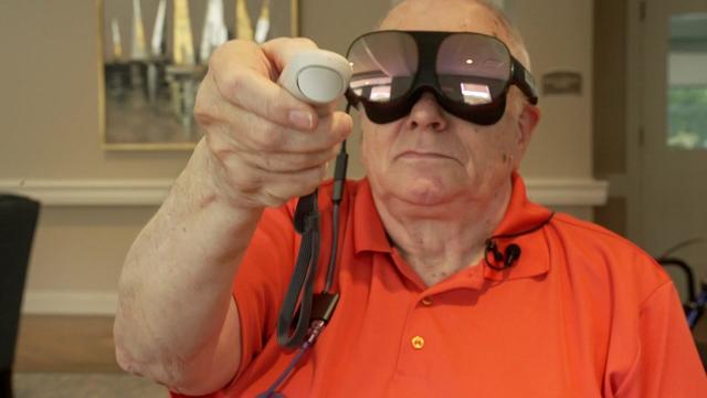 An elderly man wears VR goggles and holds a VR controller. 