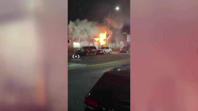 Flames and smoke pour out of a car dealership. 