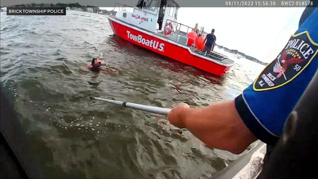 Body-worn camera shows an officer extending a boat pole to a woman in the river near a boat. 