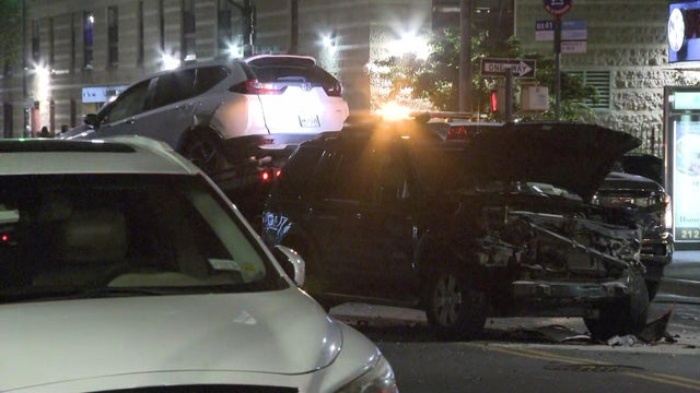 A dark-colored SUV with front end damage sits on a street. A white SUV is on a tow truck behind it. 