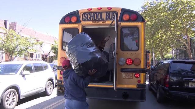 An individual hands a full garbage bag to someone standing at the back door of a school bus. 