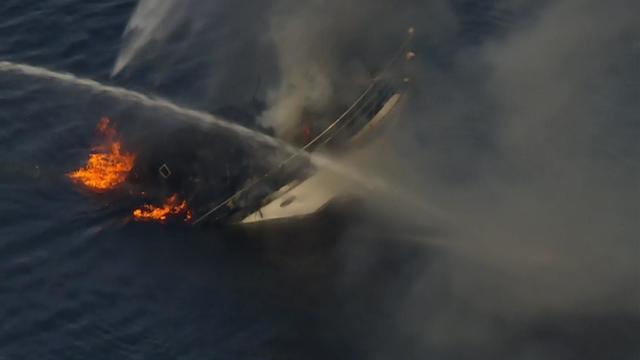 A boat on fire sinks into the water while FDNY crews spray water at it. 