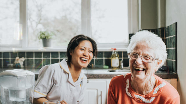 An elderly woman laughs beside a friendly young care assistant 