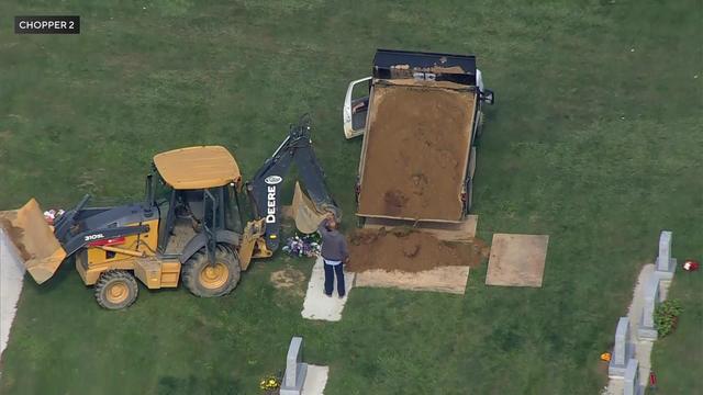 An aerial view of a piece of construction equipment filling a grave with dirt in a cemetery. 