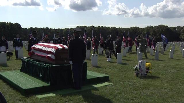 Dozens of people stand in Calverton National Cemetery as soldiers stand on either end of a flag-draped coffin. 