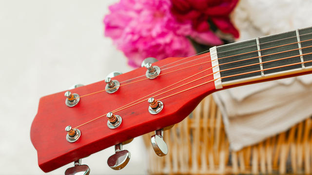brown fretboard of a six string wooden guitar and blurred multicolored peonies in a wicker basket in the background 