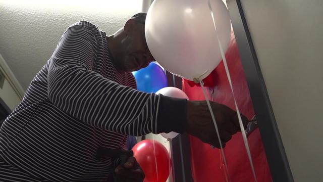 A man unlocks an apartment door. Red, white and blue balloons surround the door. 