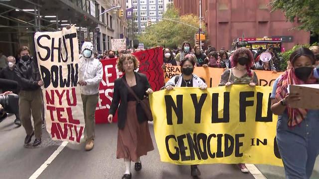 Hundreds of NYU students march down New York City streets, several carrying banners. One banner reads, "Shut down NYU Tel Aviv." 