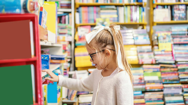 Side view of charming Caucasian blond girl with ponytail and with eyeglasses standing in bookstore and choosing what to read next. 